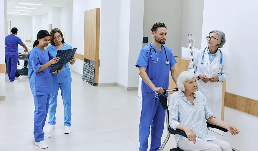 nurses and other Medical staff in the hallway of a hospital. Two nurses are speaking and looking at a clip board, one is pushing a patient in a wheelchair.